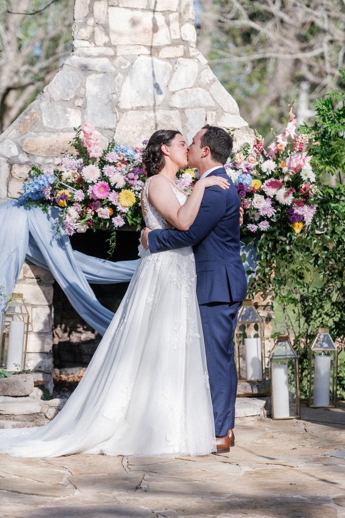 Newlyweds kissing in front of the outdoor fireplace at Pecan Springs Ranch wedding venue.
