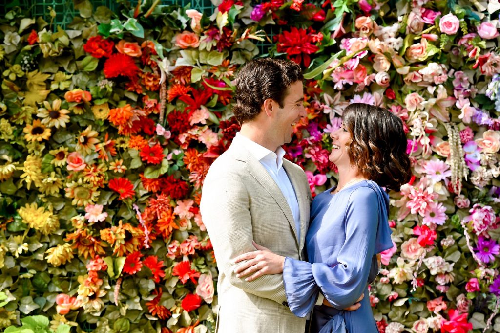 Couple smiling at each other in front of a wall of colorful flowers.