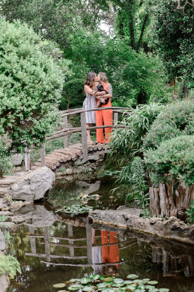 Two women kissing and holding their dog on a bridge for engagement photos at Zilker Botanical Garden.