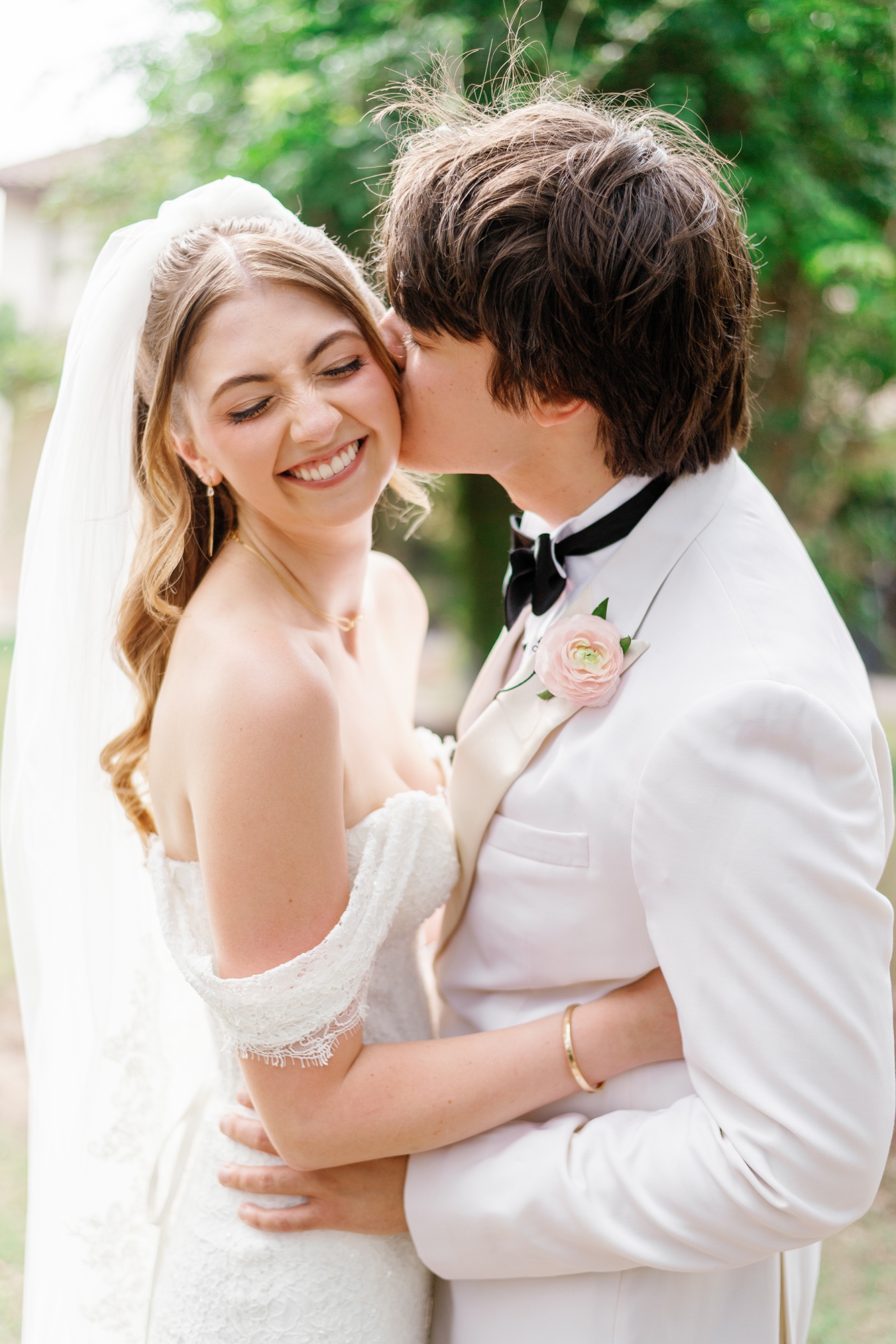 A bride smiles widely when the groom kisses her on the cheek